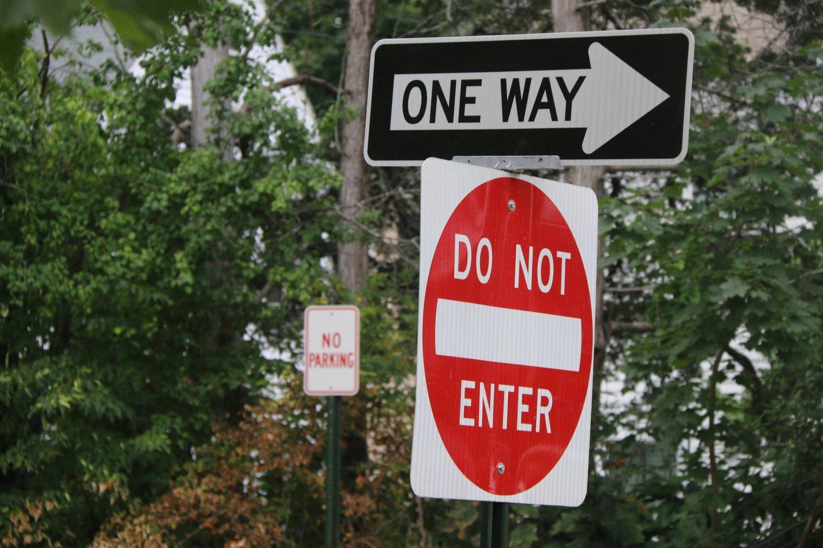 שלטי תנועה: "ONE WAY" עם חץ ימינה, "DO NOT ENTER" באדום, ו"NO PARKING" ברקע של עצים ירוקים.